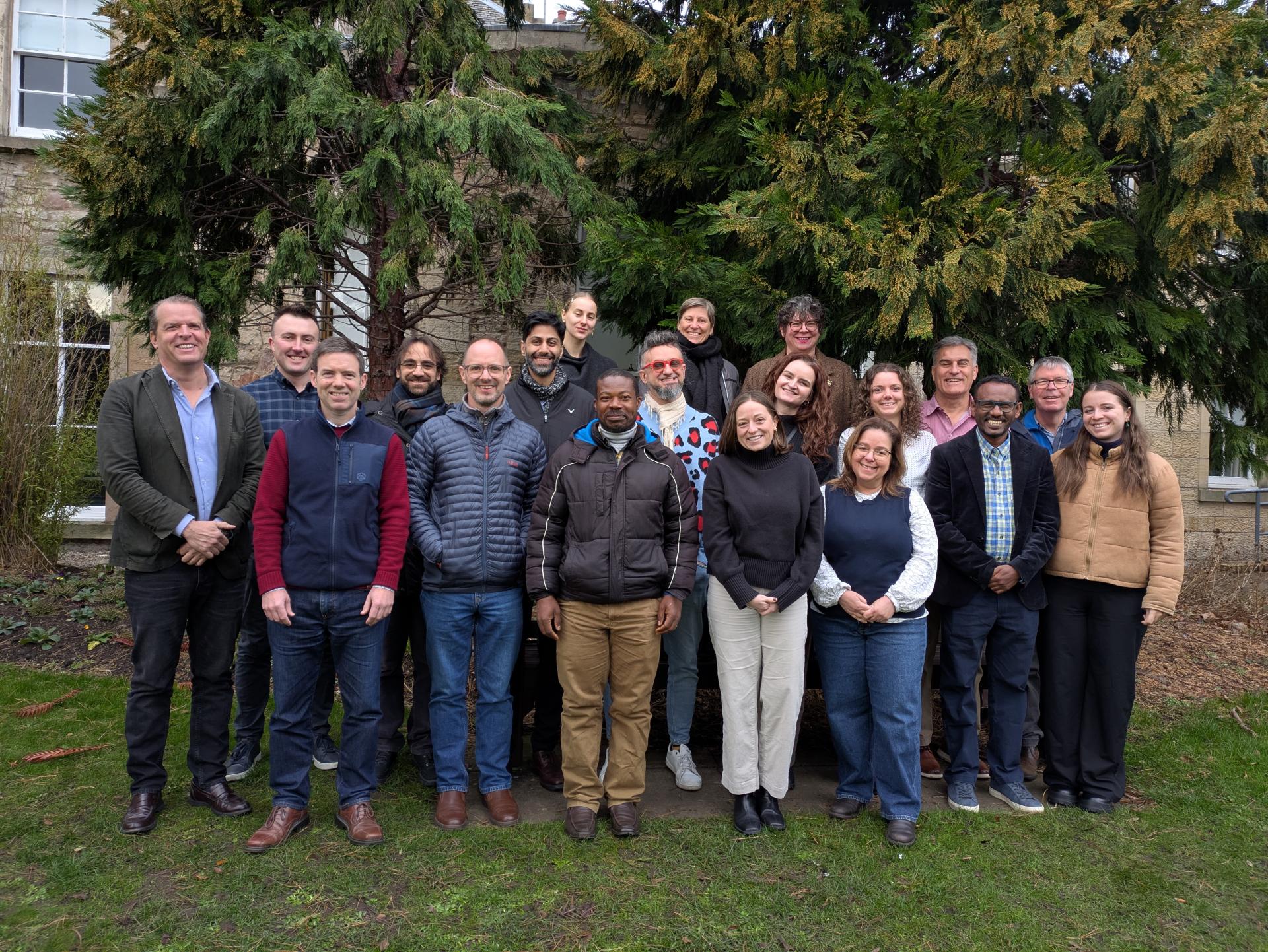 A group of adults standing outside on grass with large trees in the background.