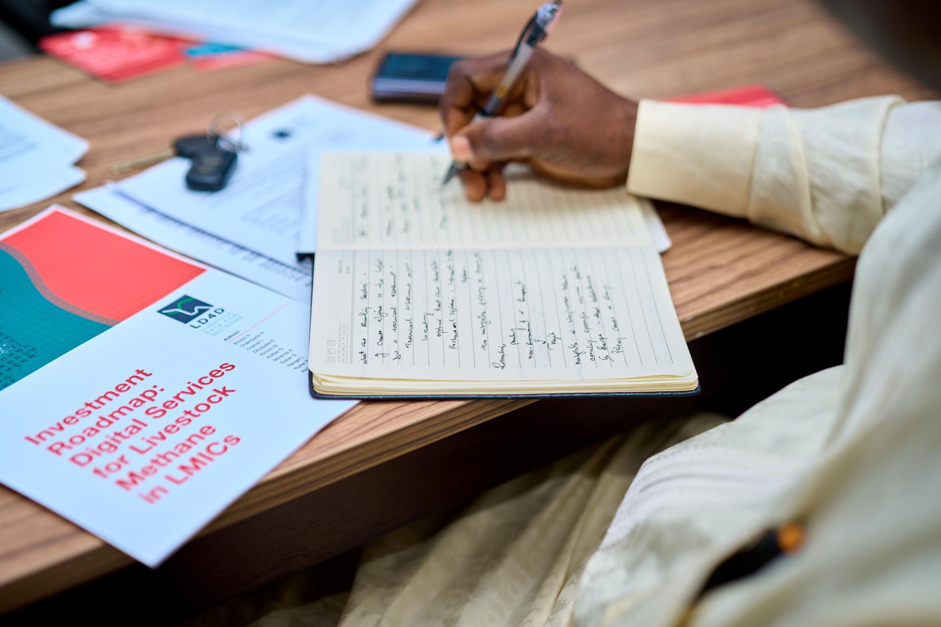 The image shows a person's hand writing in a notebook which is placed on a table. Beside the notebook are colourful documents. The face of the writer is not seen.